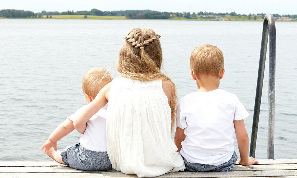 Drei Kinder sitzen auf einem Steg am See, man sieht sie von hinten