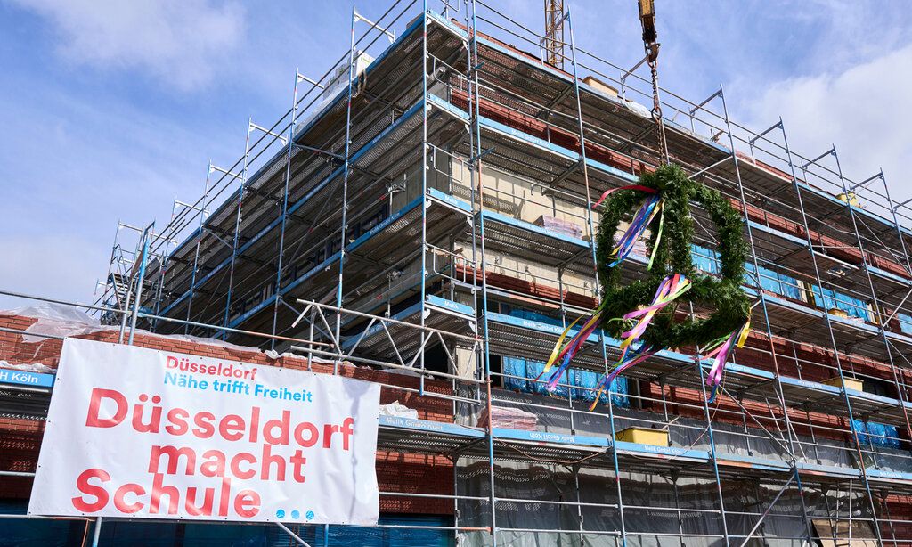 Baustelle eines Gebäudes, Banner hängt dran: Düsseldorf macht Schule, Richtkranz wird gerade befestigt