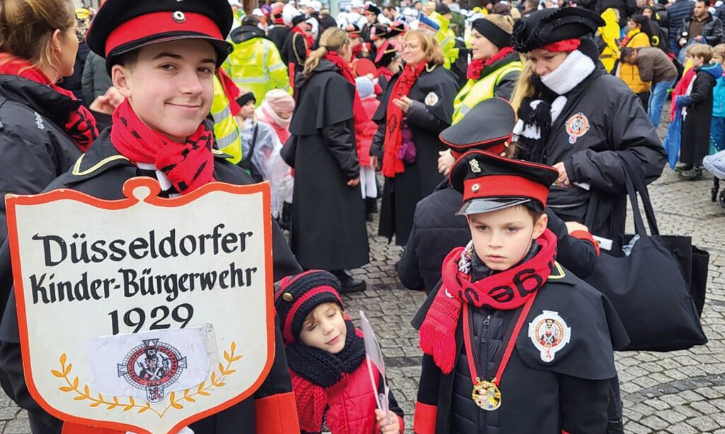 zwei Kinder und ein Jugendlicher von der Kinder-Bürgerwehr stehen in einer Menschenmenge beim Rosenmontagszug