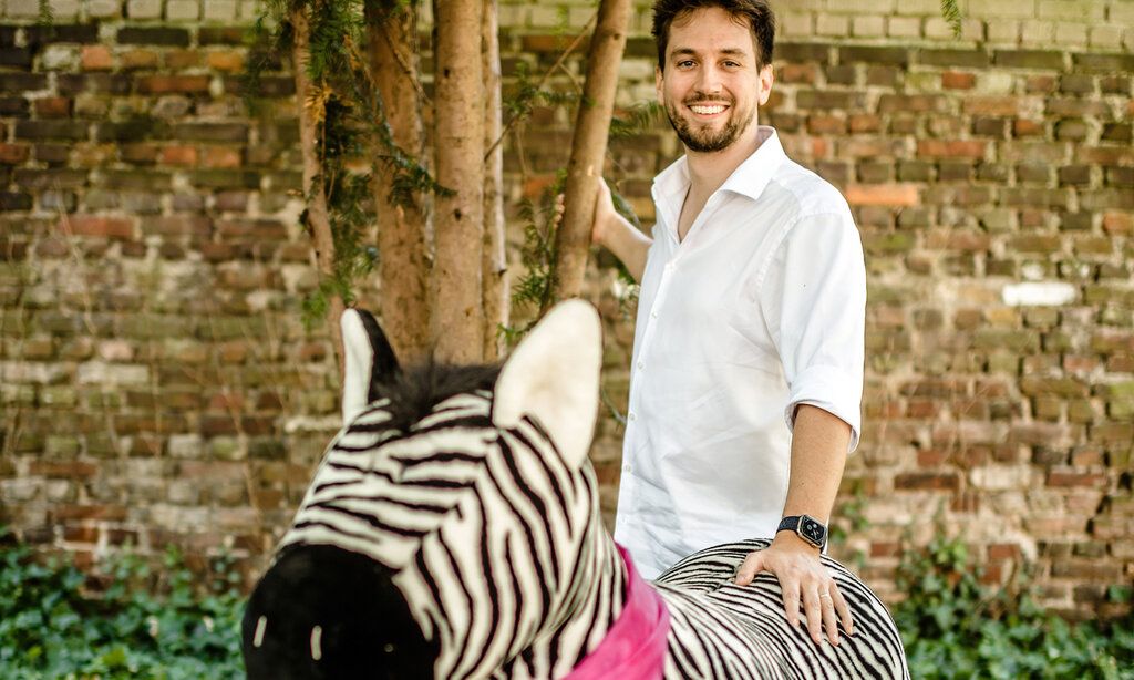 Mann im weißen Hemd mit Plüschzebra vor Baum und Mauer