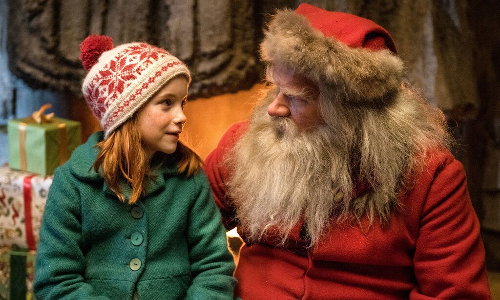 ein Mädchen mit Norweger-Mütze und grünem Mantel sitzt neben dem Weihnachtsmann und spricht mit ihm
