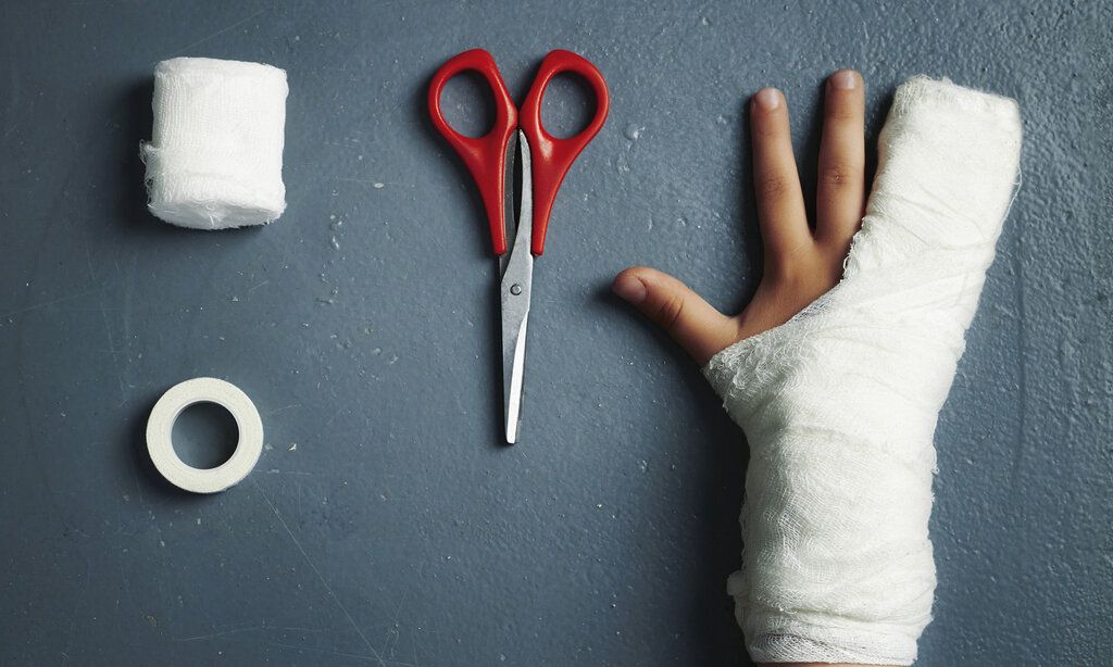 Mullbindenrolle, Kleberolle, Schere und verbundener Kinderarm von oben , dunkelgrauer Hintergrund