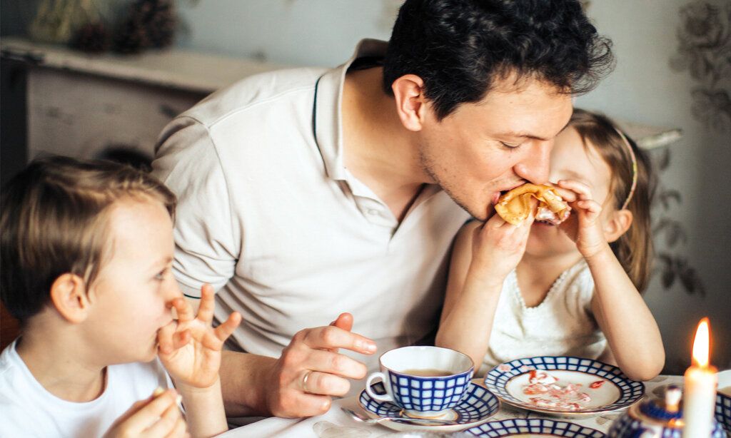 Vater sitzt mit zwei Kindern bei Tisch und beißt in den Pfannkuchen der Tochter