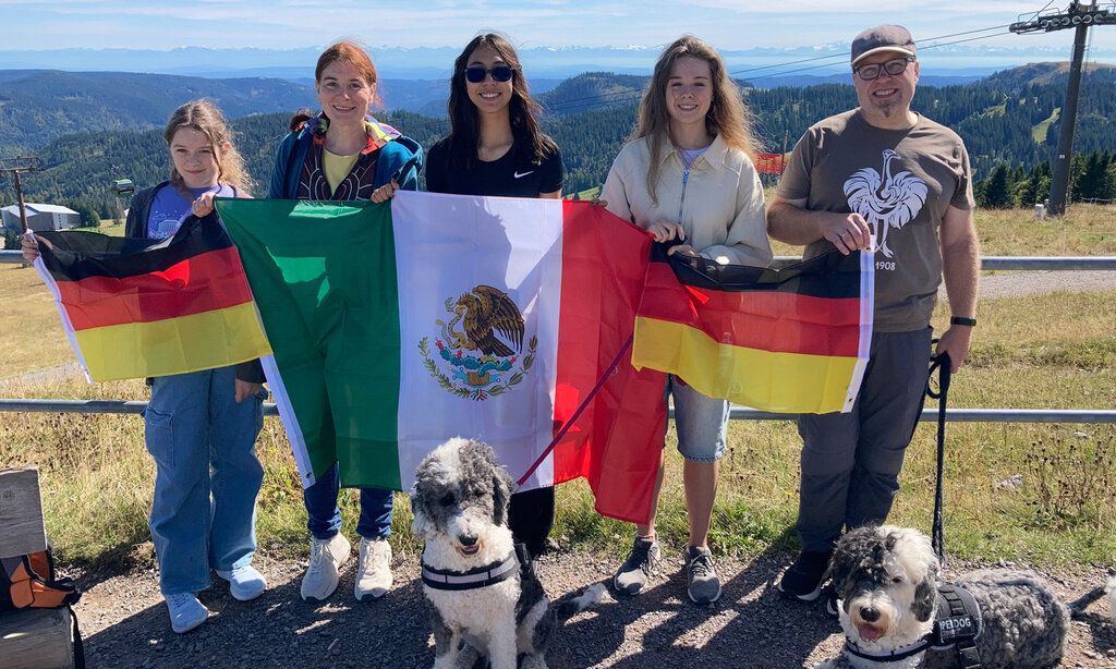 Eine Familie miteiner Austauschschülerin steht mit deutschen und mexikanischen Flaggen und zwei Hunden auf einem Berg