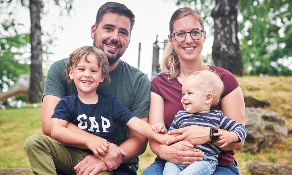 Vater und Mutter sitzen im Park, beide haben jeweils einen Sohn auf dem Schoß.