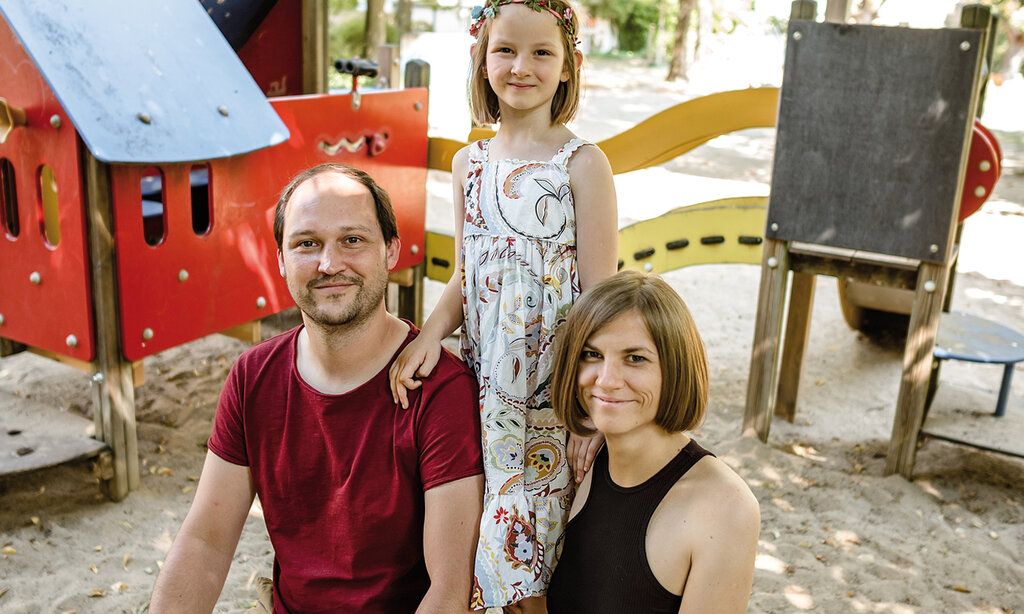 Vater und Mutter hocken im Sandkasten, Tochter steht mittig hinter ihnen