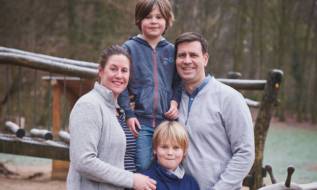 Mutter, Vater und zwei Jungs stehen zusammen auf einem Kinderspielplatz, im Hintergrund windertliche Bäume