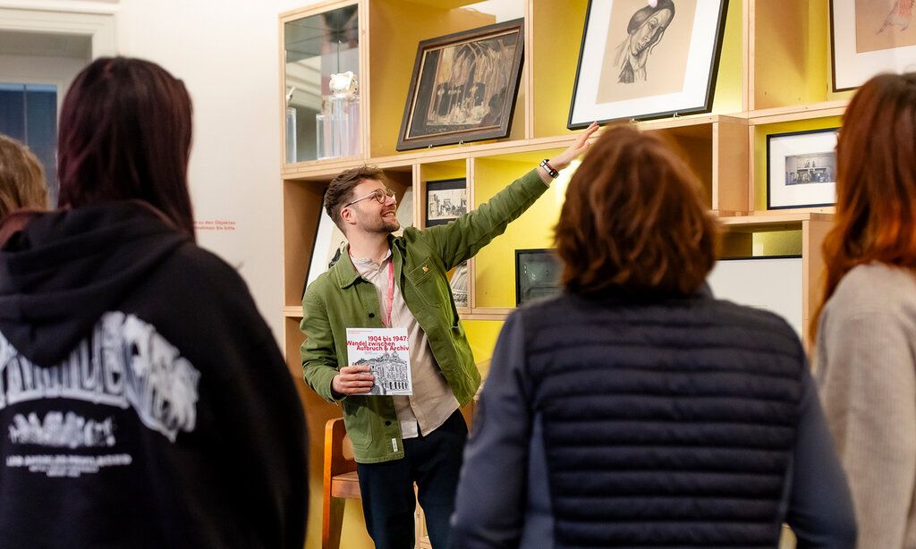 junger Mann erklärt etwas im Theatermuseum, er zeit auf ein Bild, Besucherinnen von hinten im Vordergrund