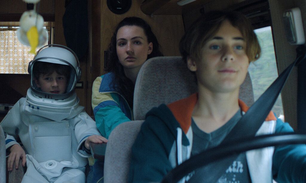 ein als Astronaut verkleidetes Kind sitzt mit einem großen Mädchen in einem Wohnmobil, im Vordergrund ein großer Junge, der das Fahrzeug fährt