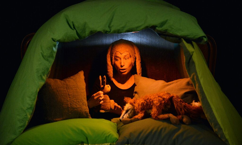 Eine Schauspielerin mit Hasenohren spielt in einer aus Kissen gebauten Höhle mit einer Hasen- und einer Fuchspuppe.