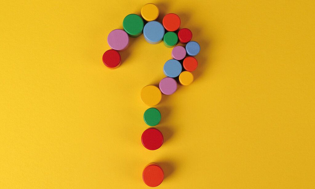 ein Fragezeichen, das aus bunten, runden Holzklötzchen gelegt ist, auf gelbem Hintergrund