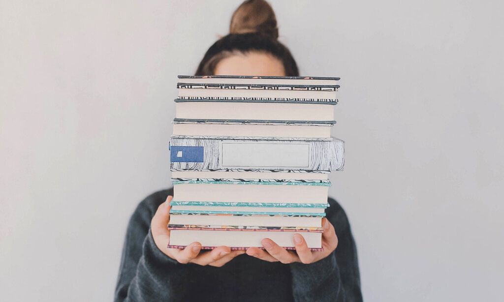 Frau hält einen Bücherstapel in die Kamera, der ihr Gesicht verdeckt