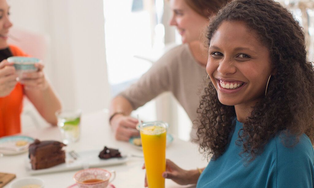 Frau sitzt an einem Esstisch mit einem Glas Orangensaft, im Hintergrund zwei Frauen, die sich unterhalten