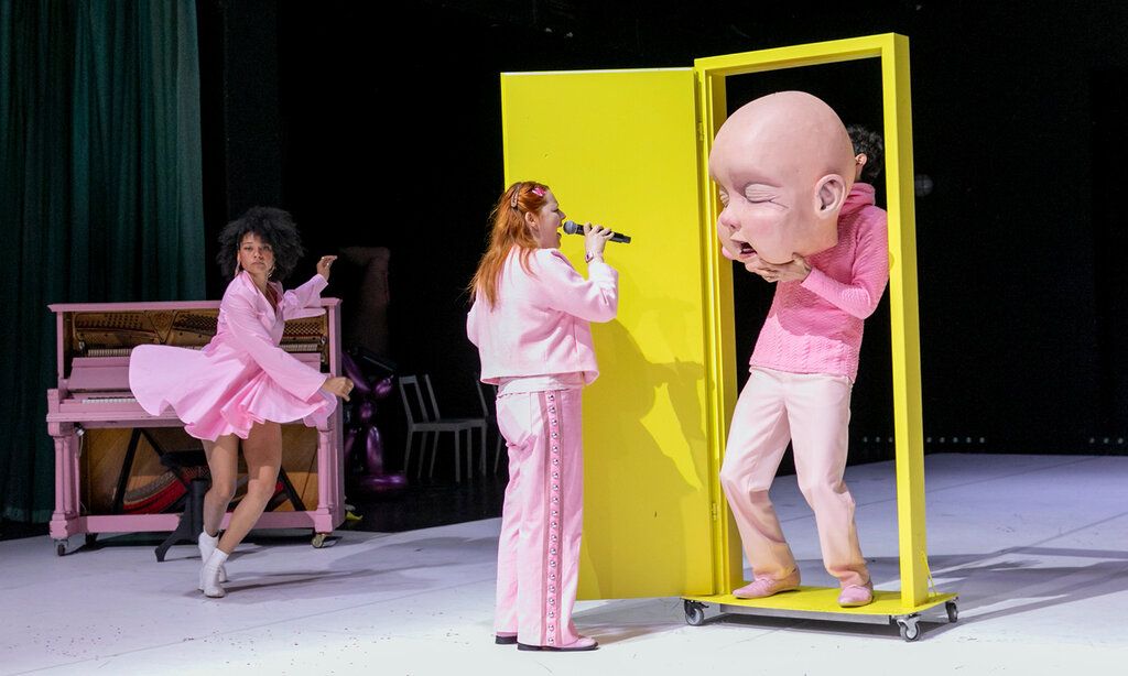 drei Schauspieler:innen in rosa Kleidung auf der Bühne, eine gelbe Tür auf der Bühne, ein Schauspieler hält sich eine große Baby-Maske vors Gesicht.