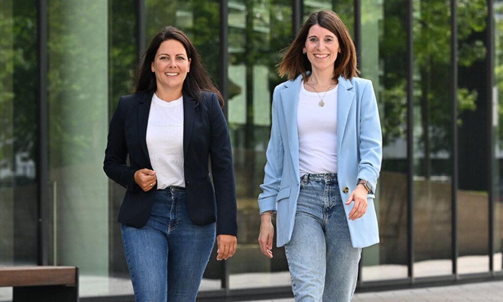 Zwei Frauen, Anika Schmidt und Lena Pieper, im Business-Outfit laufen nebeneinader her, im Hintergrund spiegelnde Scheiben eines Bürohauses