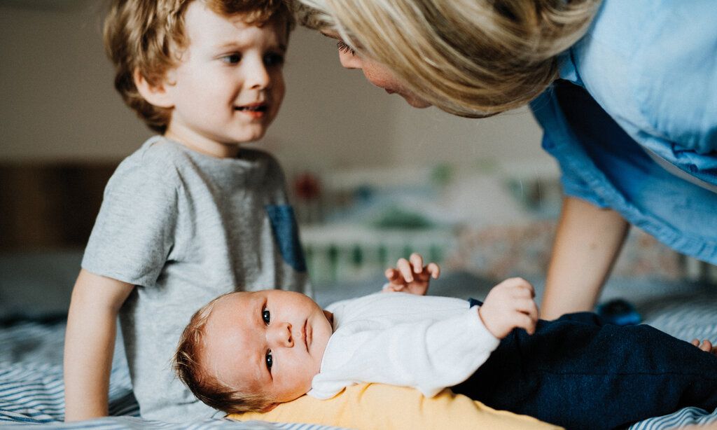 Baby liegt auf einer Decke und guckt in die Kamera, Bruder sitzt dabei und wirkt unzufrieden, Mutter spricht mit ihm