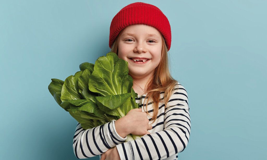 Mädchen mit Ringel-Shirt und roter Mütze hält Salat im Arm, vor blauem Hintergrund