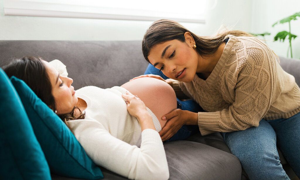 Schwangere Frau liegt auf einem Sofa und wird von einer Hebamme untersucht.