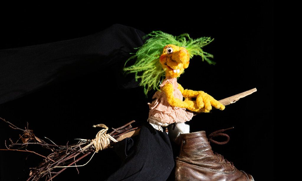 Die Puppe Hexe Hilda reitet vor schwarzem Hintergrund auf ihrem Besen.