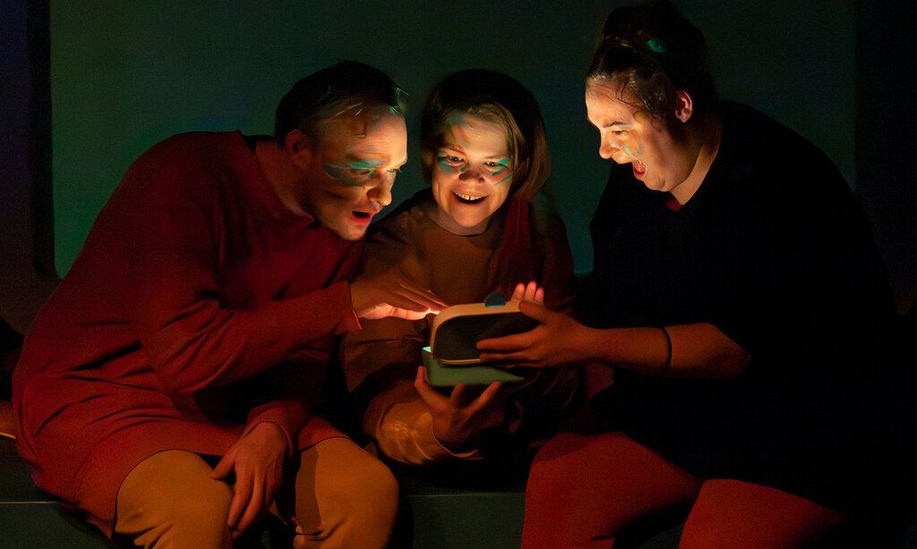 ein Schauspieler  und zwei Schauspielerinnen sitzen im Dunklen zusammen und gucken auf etwas leuchtendes