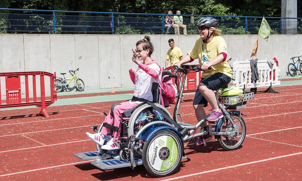 Special Petit Départ für Kinder mit Handicap im Rather Waldstadion, Frau fährt mit speziellem Rad, auf dem ein Rollstuhl installiert ist, auf dem ein Mädchen sitzt, auf einer Aschebahn