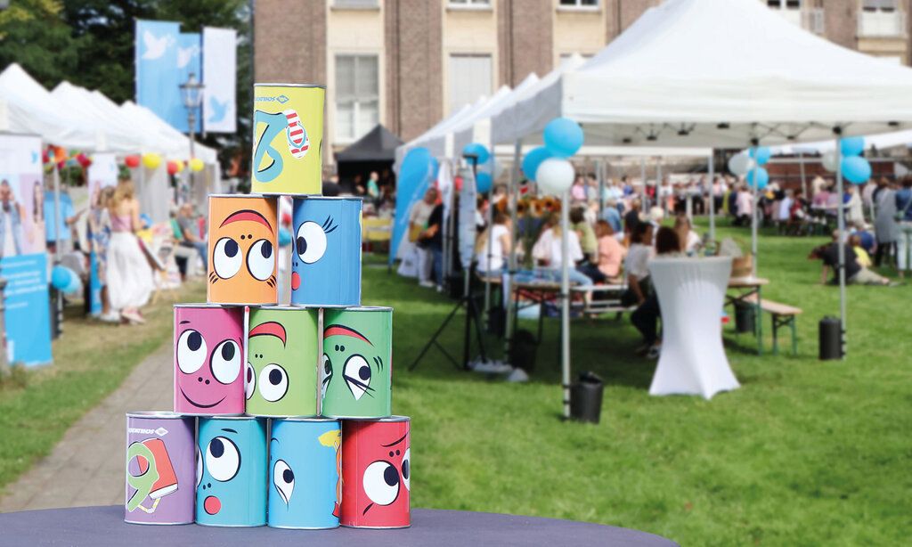 Jahresfest inder Kaiserswerther Diakonie, Biertische und Pavillons im Garten der Diakonie mit Besucher:innen, im Vordergrund gestapelte Blechdosen mit lustigen Gesichtern