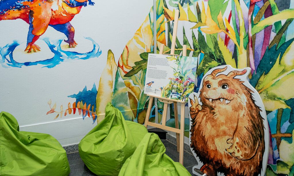 Ecke eines Ausstellungsraumes im Oberschlesischen Landesmuseum mit grünen Sitzsäcken, einem Aufsteller in Form eines kleinen Monsters und eine Staffelei, auf der ein aufgeschlagenes Buch liegt