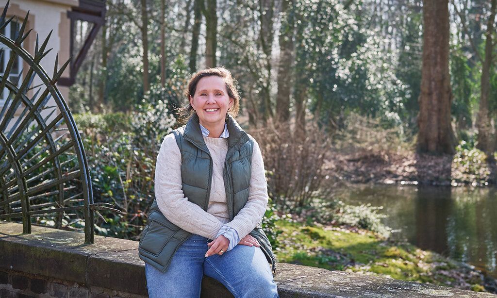 Johanna Adolphs-Kenning sitzt auf einem Mäuerchen vor dem Schloss Eller und dem Schlosspark
