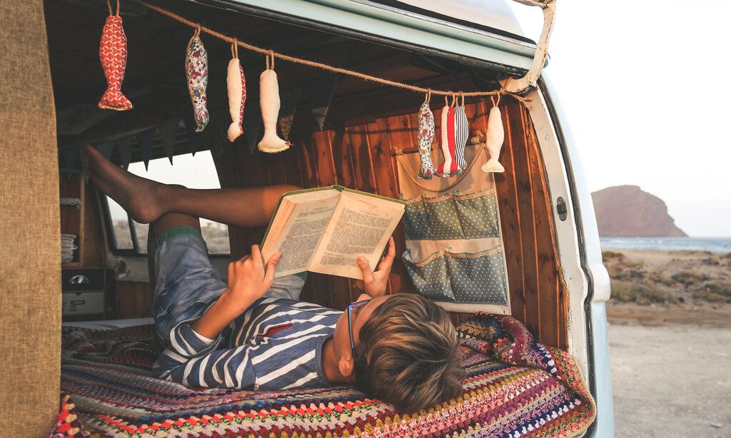 Junge liest in einem Bully ein Buch, das Auto steht am Strand