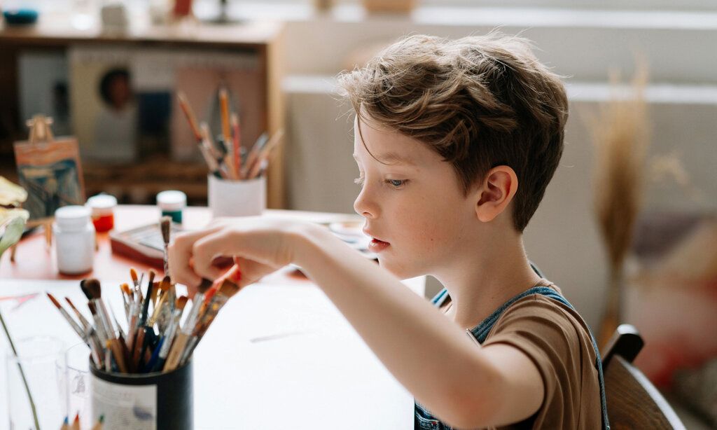 Junge sitzt am Tisch mit Malutensilien und nimmt einen Pinsel aus einem Becher mit Pinseln
