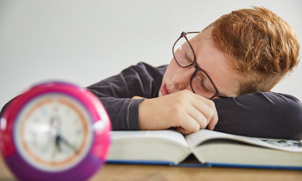 ein Junge schläft auf einem Buch, im Vordergrund steht ein Wecker