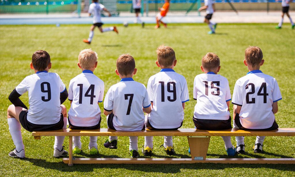 Jungs einer Fußballmannschaft sitzen auf dem Fußballplatz auf einer Bank, man sieht sie von hinten
