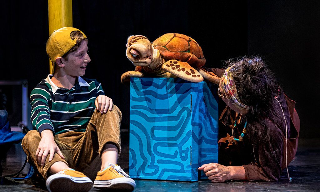 Junge sitzt auf dem Boden und spricht mit Schildkrötenpuppe daneben sitzt die Puppenspielerin