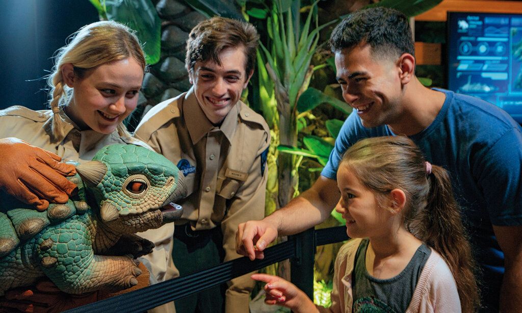 Zwei Museumsmitarbeiter:innen halten eine Dinosaurierfigur, ein Mädchen und sein Vater gucken sich die an