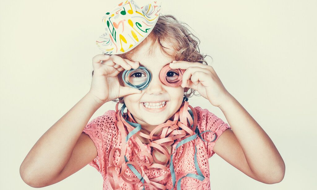 Mädchen mit Karnevallshut hält sich Luftschlangen vor die Augen und lacht, Hintergrund hellgelb