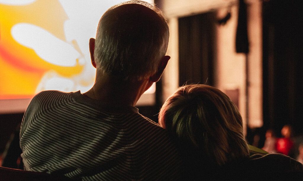 erwachsener Mann und Kind im Kino, von hinten fotografiert, Kopf des Kindes lehnt an der Schulter des Mannes, im Hintergund die leuchtende Leinwand