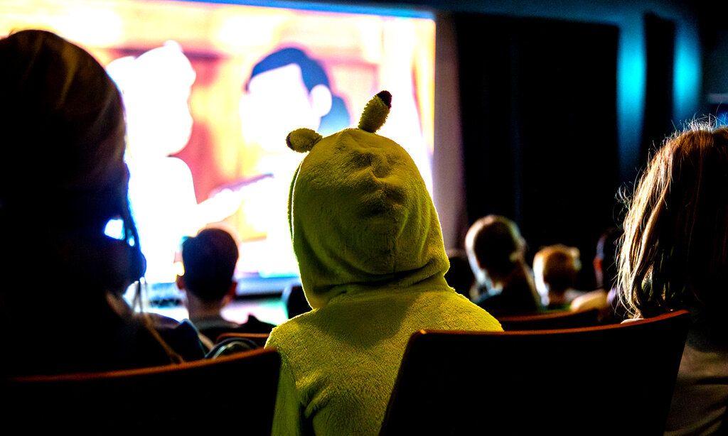 ein Kind sitzt beim Kinderkinofest im Kinosaal, es trägt mit einen Kapuzenpilli mit Ohren, man sieht das Kind von hinten, im Hintergrund sieht man die Kinoleinwand