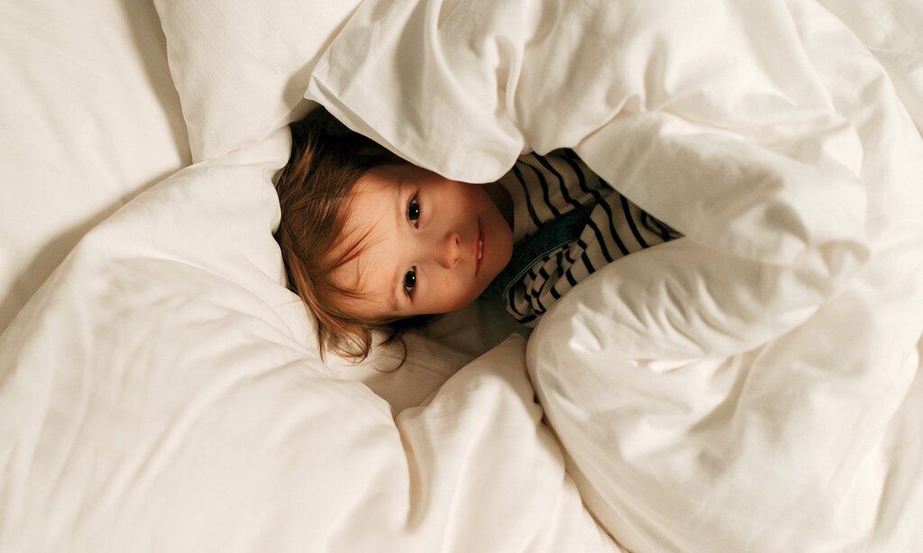 Kind liegt im Bett und guckt unter der Bettdecke hervor