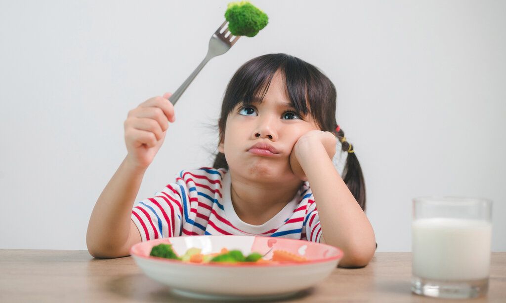 kleines Mädchen hat ein Brokkoliröschen auf der Gabel und guckt es trotzig an