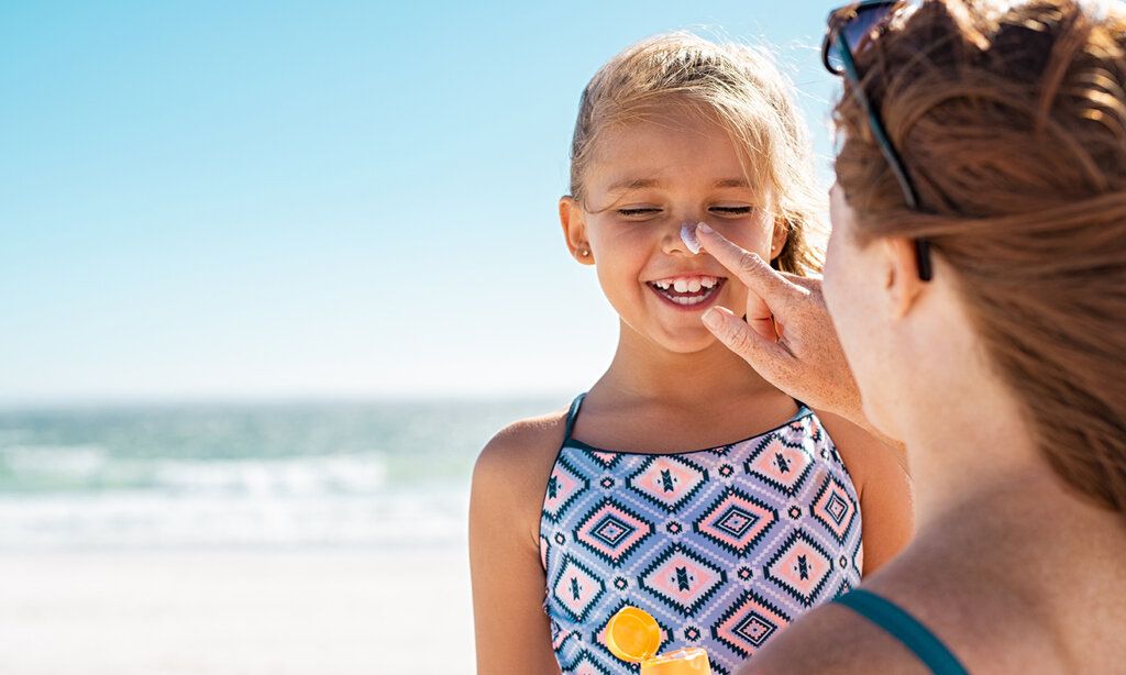 Ein Mädchen wird von seiner Mutter am Strand mit Sonnencreme eingecremt, im Hintergrund Meer
