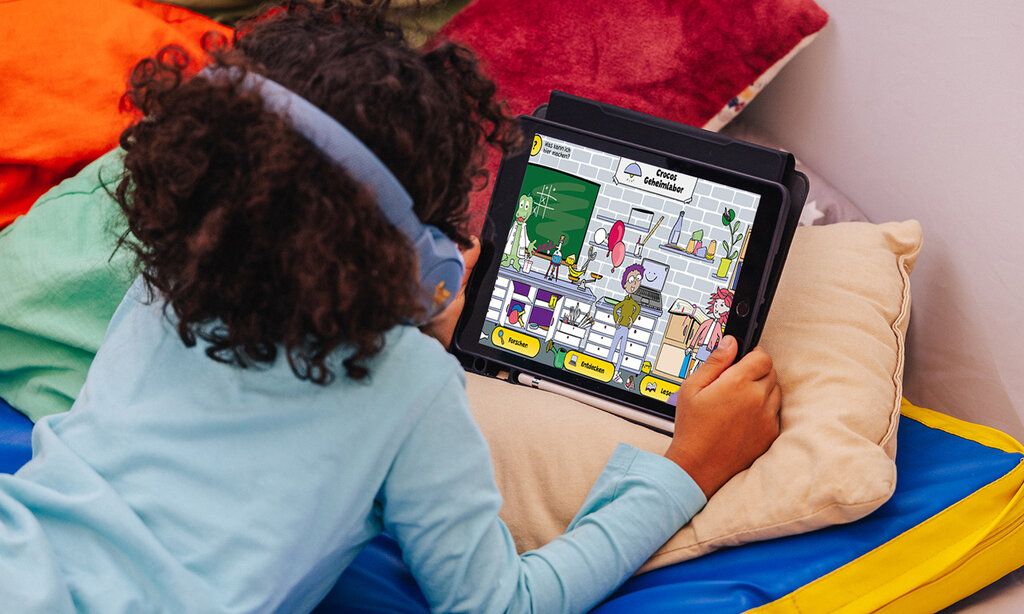 Kind liegt mit Tablet auf einer Matratze und betrachtet Kinderwebsite Crocos Geheimlabor