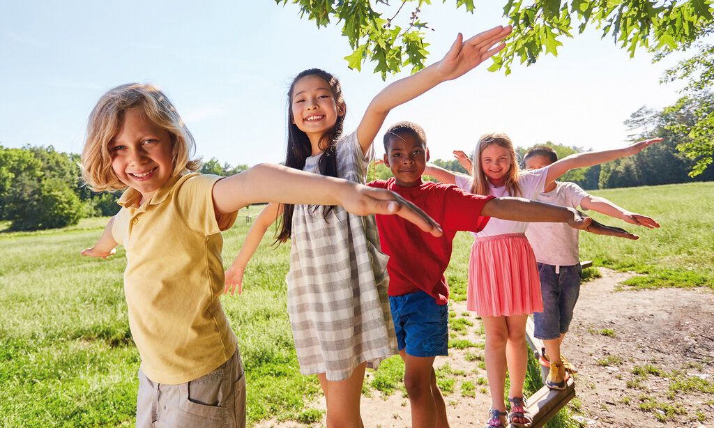 Fünf Kinder balancieren auf einem Holzbalken, im Hintergrund eine Wiese und Bäume