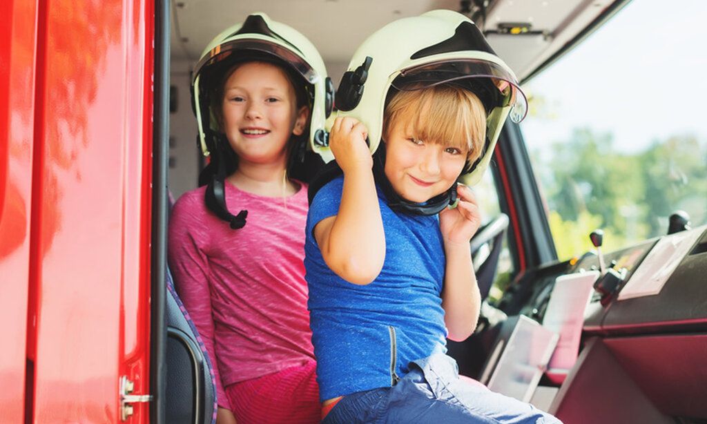 Zwei Kinder sitzen im Führerhaus eines Feuerwehrwagens, beide tragen Helme und gucken in die Kamera