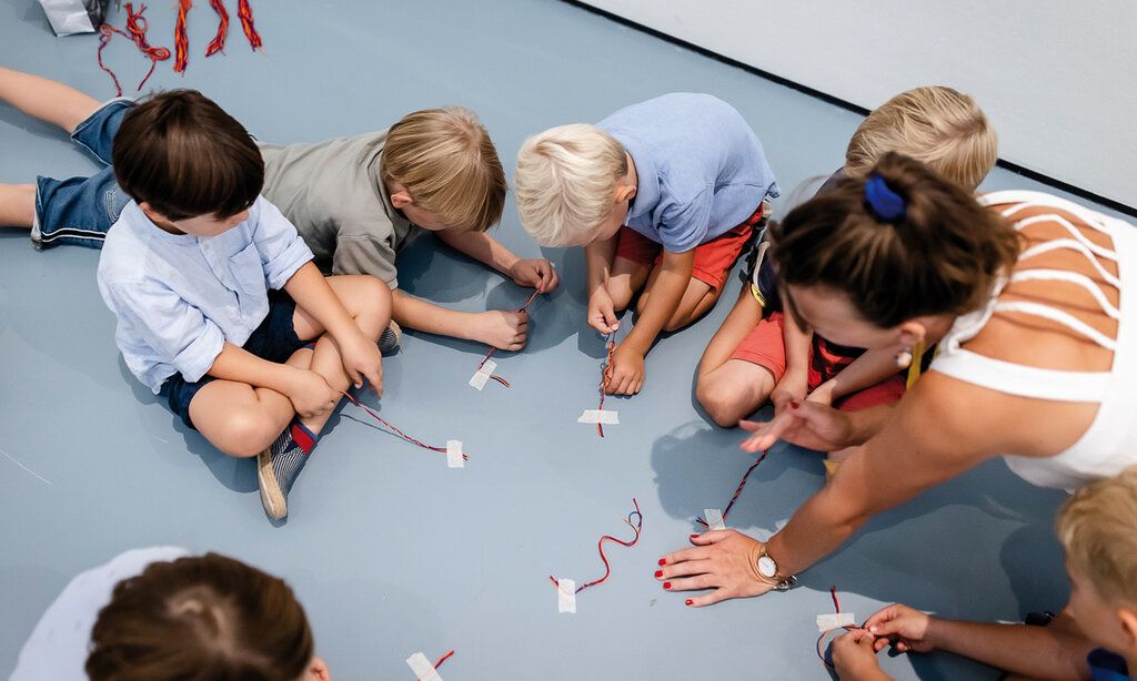 Meherere Kinder sitzen auf einem blauen Boden und basteln mit Drähten, eine erwachsene Frau hilft