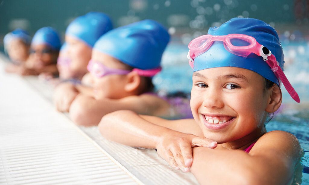 mehrere Kinder im Schwimmbecken, halten sich am Beckenrand fest, alle tragen die gleichen Badekappen und Chlorbrillen, das vordere Mädchen lacht in die Kamera