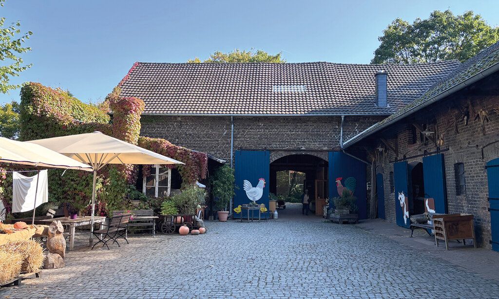Kinderbauernhof Neuss, Blick auf Bauernhaus mit Toren, Vorplatz mit Heuballen, Sonnenschirmen, Sitzgelegenheit und Kürbissen