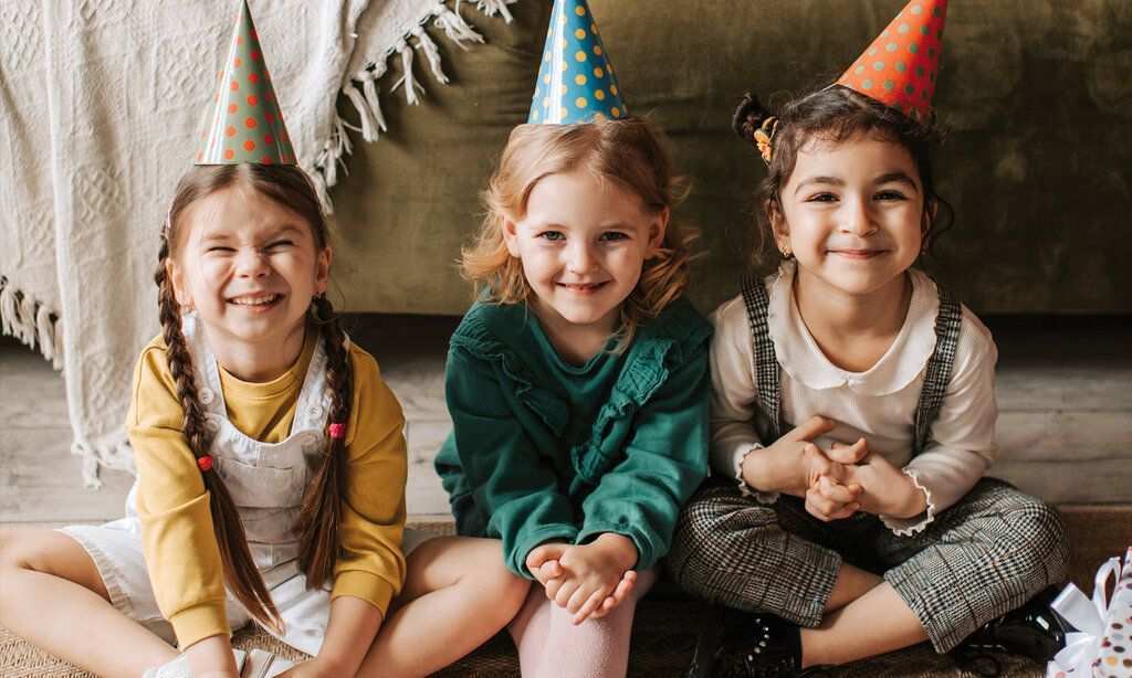 drei Mädchen mit Partyhütchen sitzen lachend vorm Sofa auf dem Boden