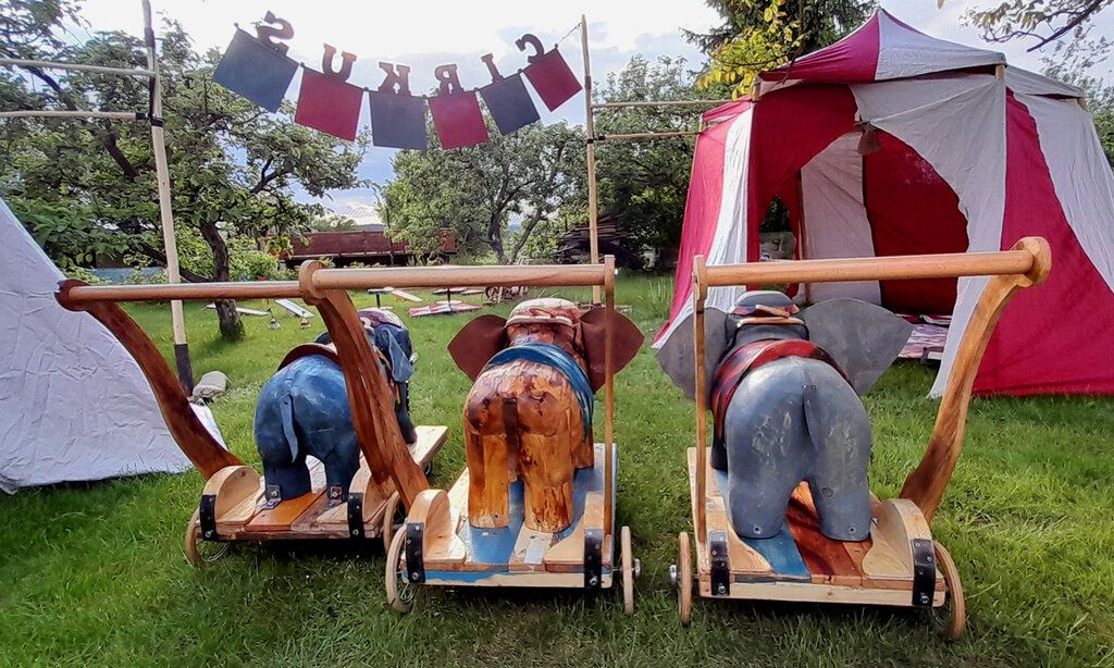 eine Wiese mit kleinen Zelten und Elefantenfiguren auf Rädern von hinten