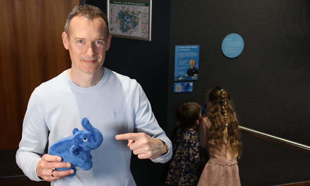 ein mann hält einen Plüschelefant in der Hand und zeigt an ihn, im Hintergrund: zwei Kinder von hinten