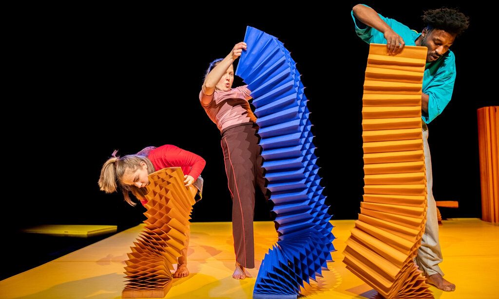 drei Schauspieler:innen spielen auf der Bühne mit überdiemensionalen, bunten, zieharmonikaähnlichen Objekten